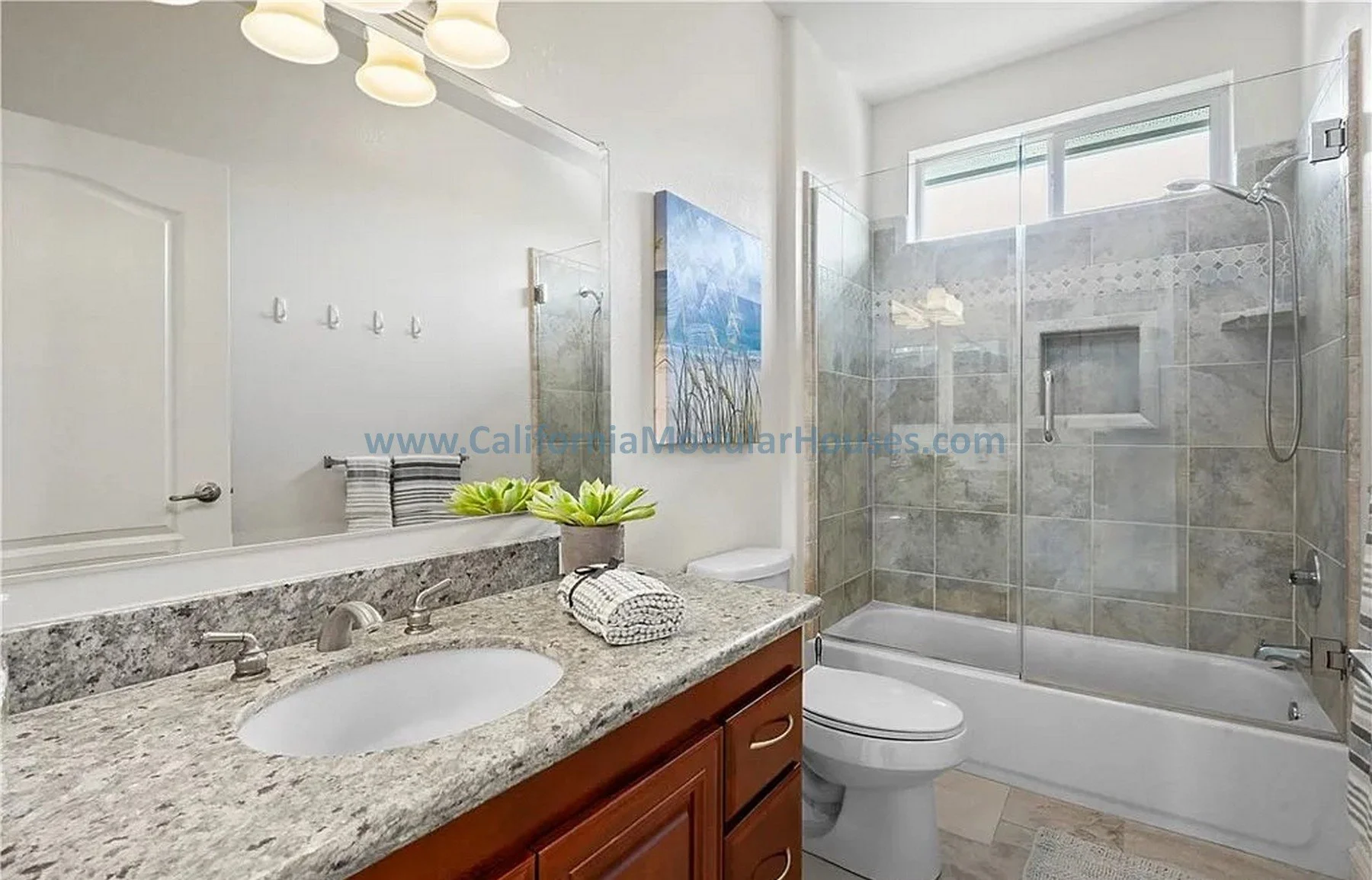 Bathroom with granite countertop vanity large mirror, shower with glass door, gray tiled walls, window, and toilet.  California Modular Houses. Pre fab home.  Modular homes for California.  Casas prefabricadas.  Southern California.  