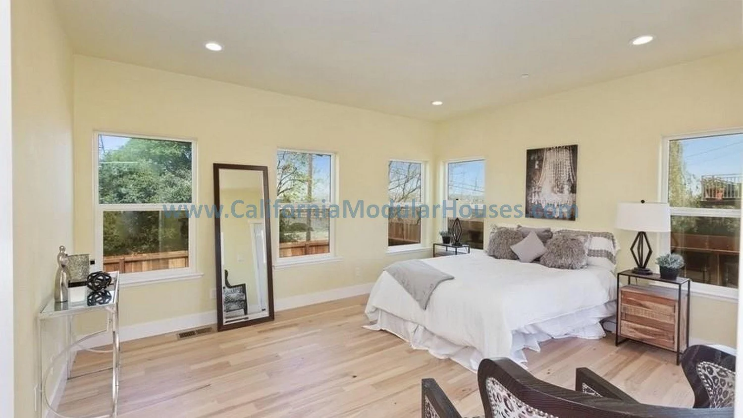 Bedroom with yellow walls, hardwood floors, a white bed with pillows, nightstand, table lamp, and a full-length mirror leaning against the wall, with three windows showing trees outside.  Modular house interior.  