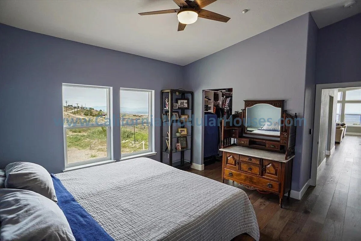 A bedroom with light blue walls, a bed with white bedding and pillows, two windows showing a hill outside, a ceiling fan, a black bookshelf with pictures and books, and a vintage wooden vanity with a mirror.