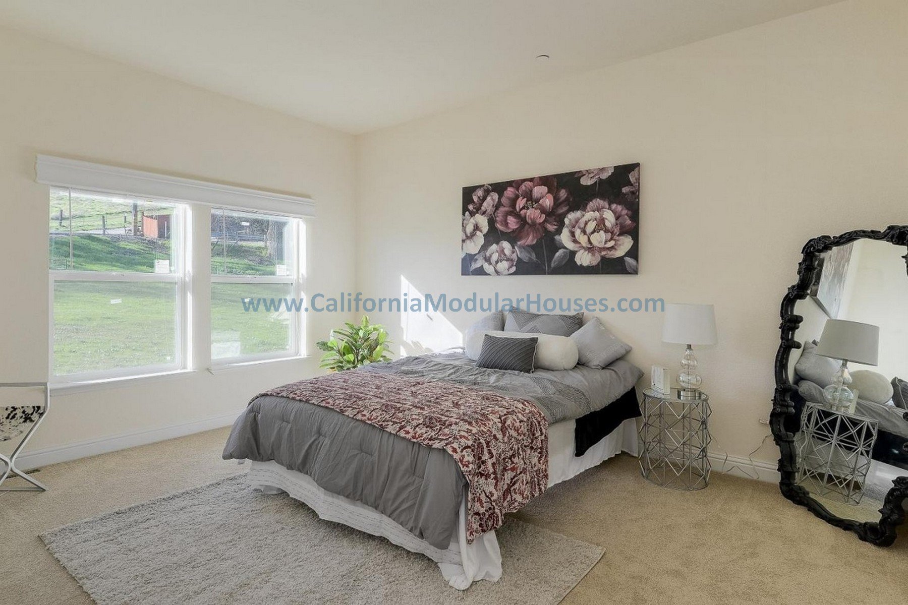 Bedroom with large window, bed with gray bedding and decorative pillows, black framed mirror, white lamps, floral artwork, potted plant, beige carpet, white walls.  San Jose Modular Home, Prefab Home California,