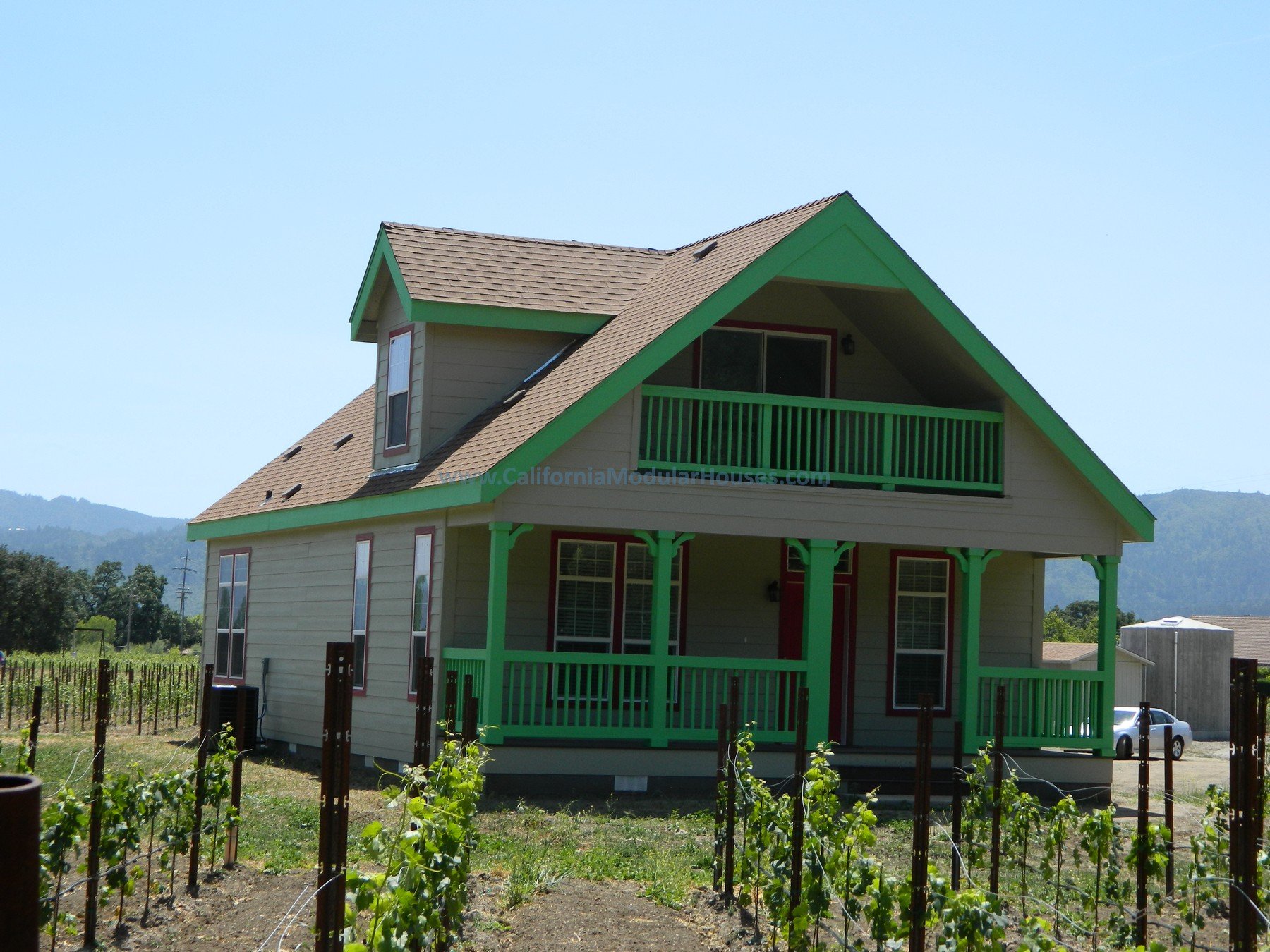 Two-story house with beige exterior and green trim, surrounded by a garden with young plants, under a clear blue sky, with mountains in the background.