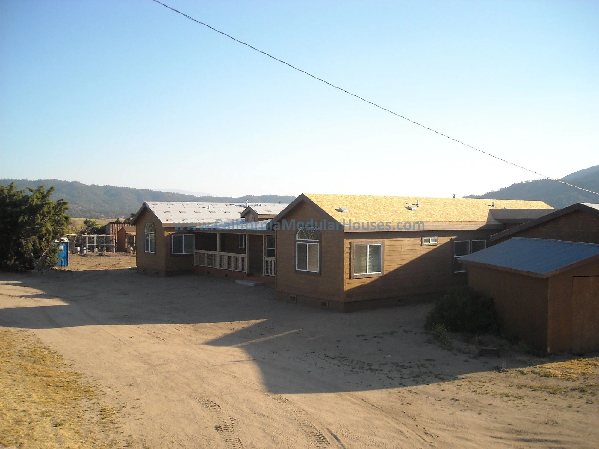 Single-story wooden modular house with a gabled roof, some windows, and small porch, situated in a rural area with mountains in the background.