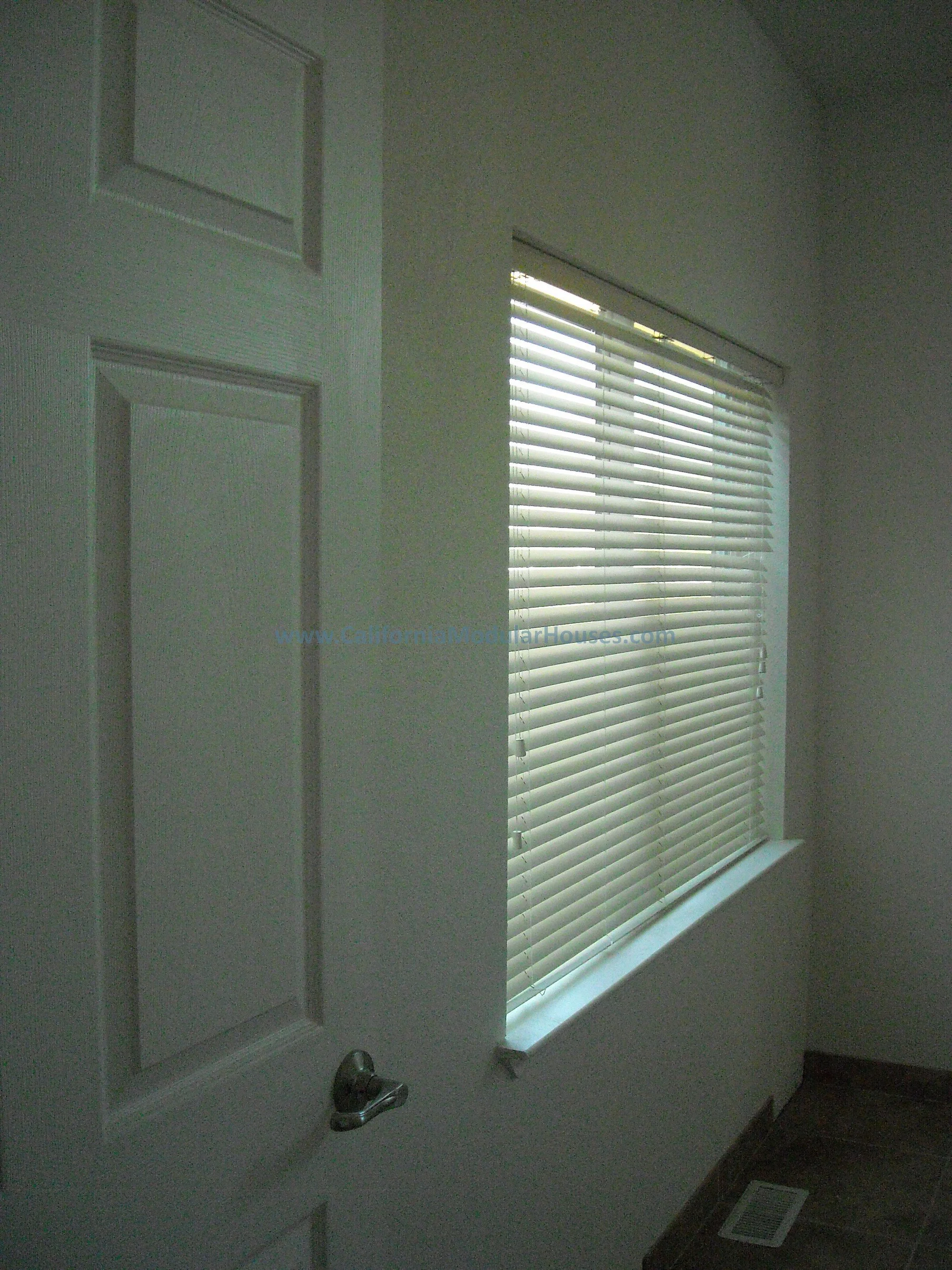 Interior of a room with a closed white door on the left and a window with closed horizontal blinds on the right.  This modular home has many nice, large windows for plenty of living.  