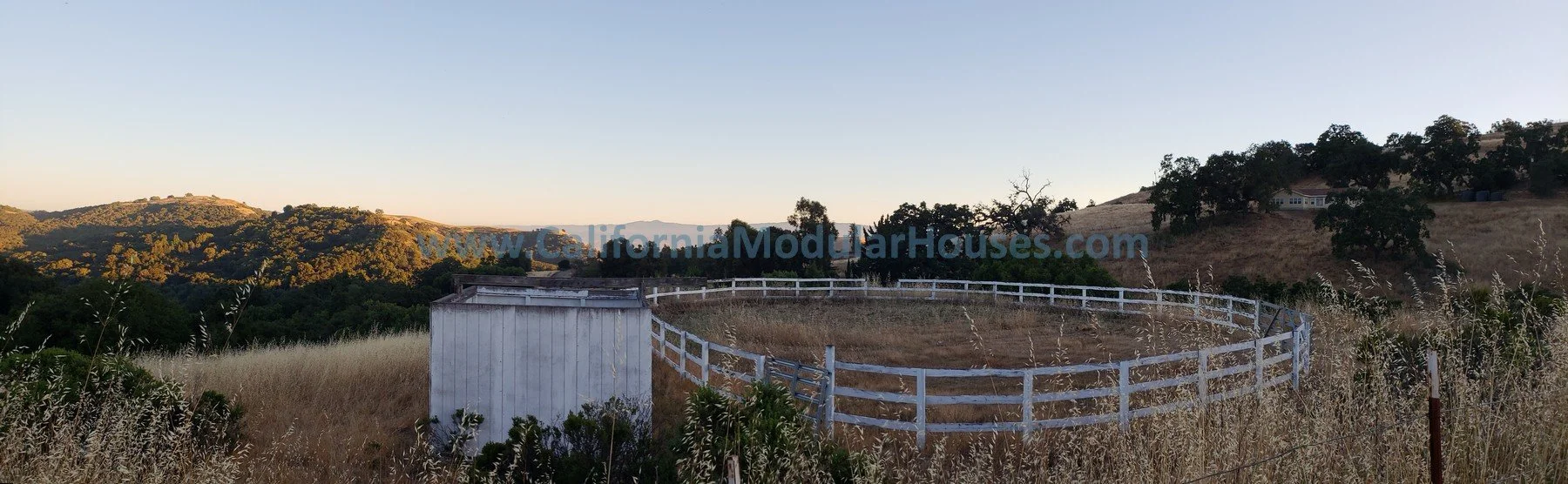 A rural landscape with rolling hills, dry grass, scattered trees, and a white wooden fence enclosing a small structure, possibly a shed, with a body of water nearby, under a clear sky at sunset. Modular Homes,