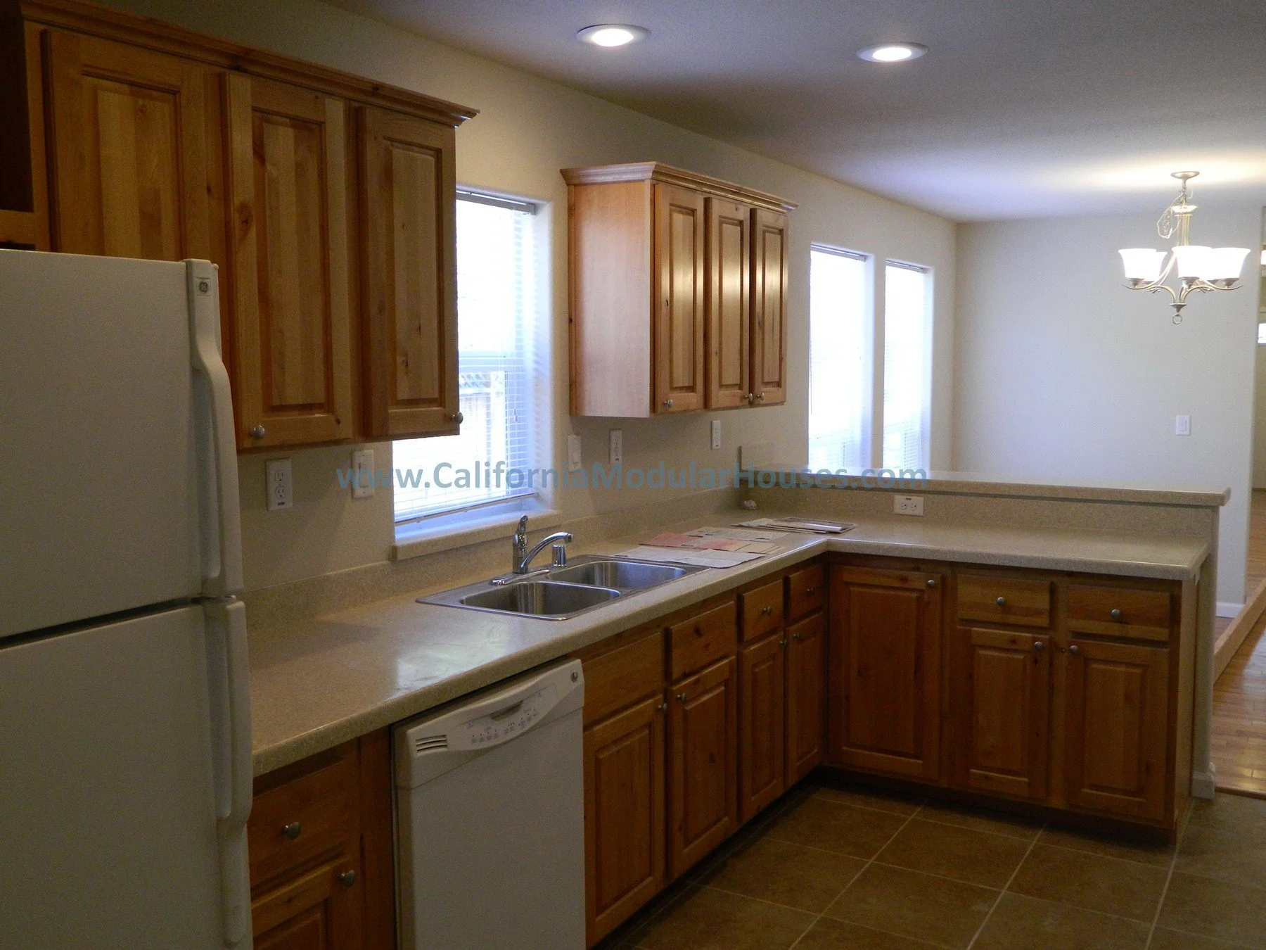Kitchen with wooden cabinets, white refrigerator, dishwasher, double sink, and laminate countertops, illuminated by ceiling lights and natural light from windows.