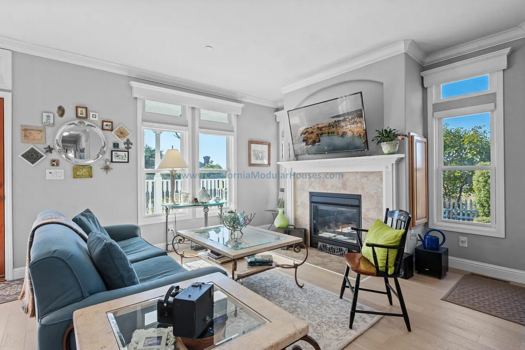 Living room with a blue sofa, glass coffee table, fireplace, and large windows showing outside greenery. Modular prefab house. California. Cape Cod home with attic finished on-site.