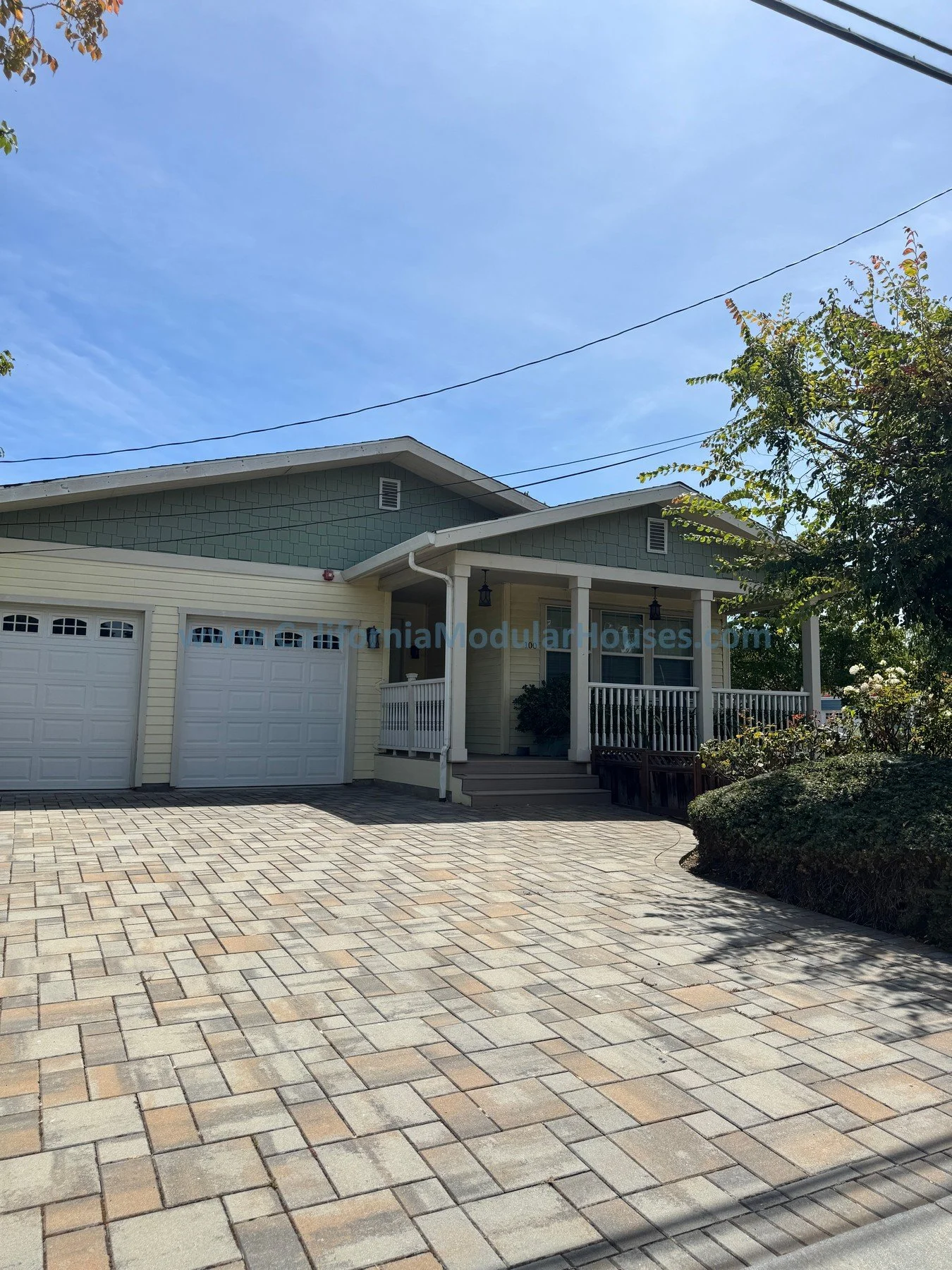 A two-story house with a yellow exterior, gray shingles on the upper section, a front porch with white railing, and a double garage with white doors. The driveway is paved with multicolored bricks. There are trees and bushes around the house, under a