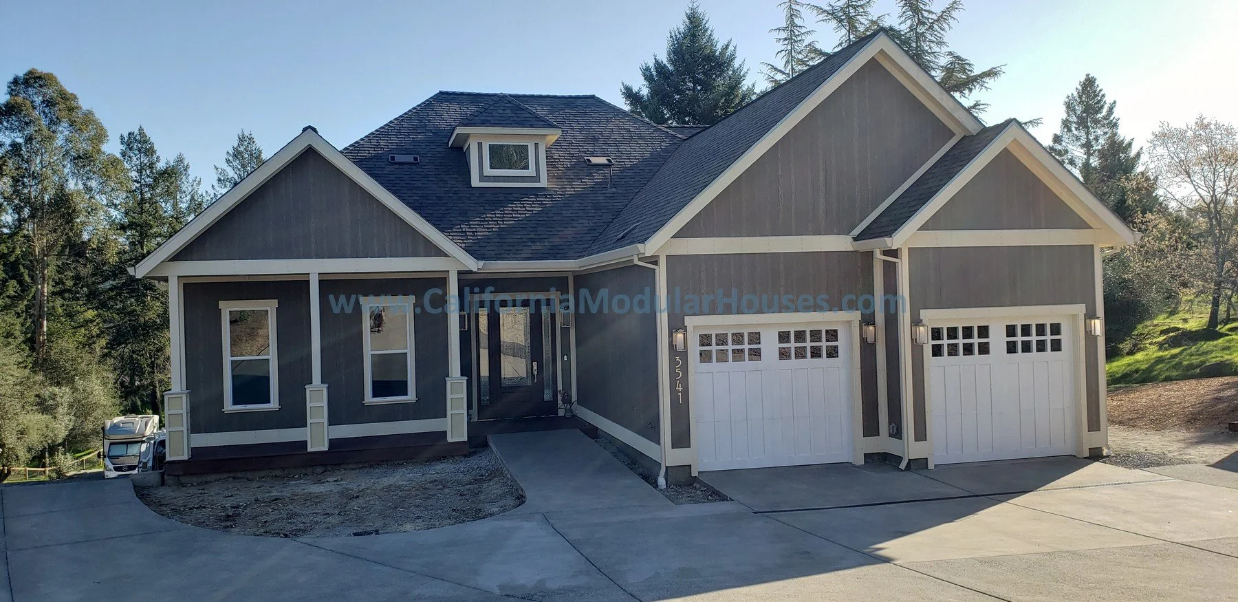 Modern gray house with white trim, two garage doors, and a front porch, surrounded by trees under a clear sky.  Santa Rosa, CA.  Sonoma County.  Modular Homes Northern California, Prefab Houses, Prefab Home California,