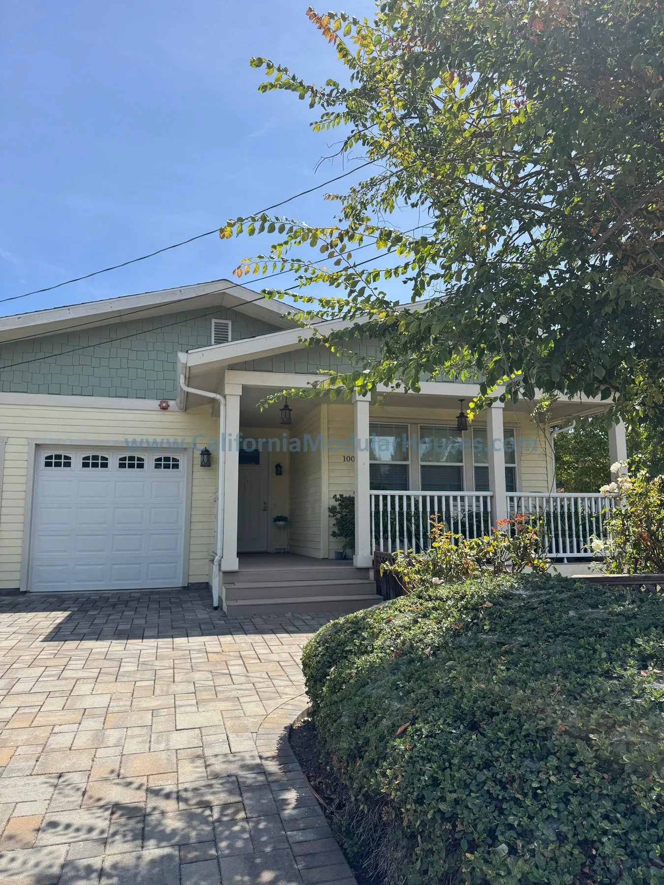Front view of a modern house with a white garage door, light yellow exterior walls, and a porch with white railings and columns. There is a paved driveway, some greenery, bushes, and a tree on the right side. The house has a gabled roof with green sh