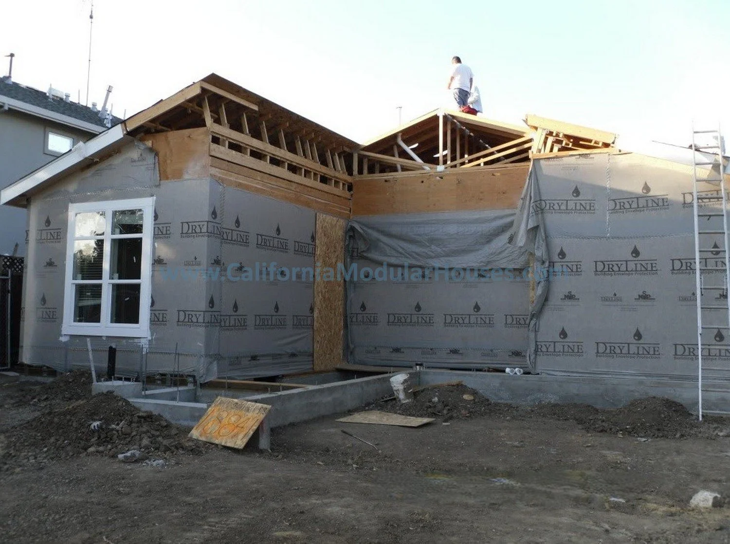 Construction site of a house with partially built walls, a window, and workers on the roof framing stage.