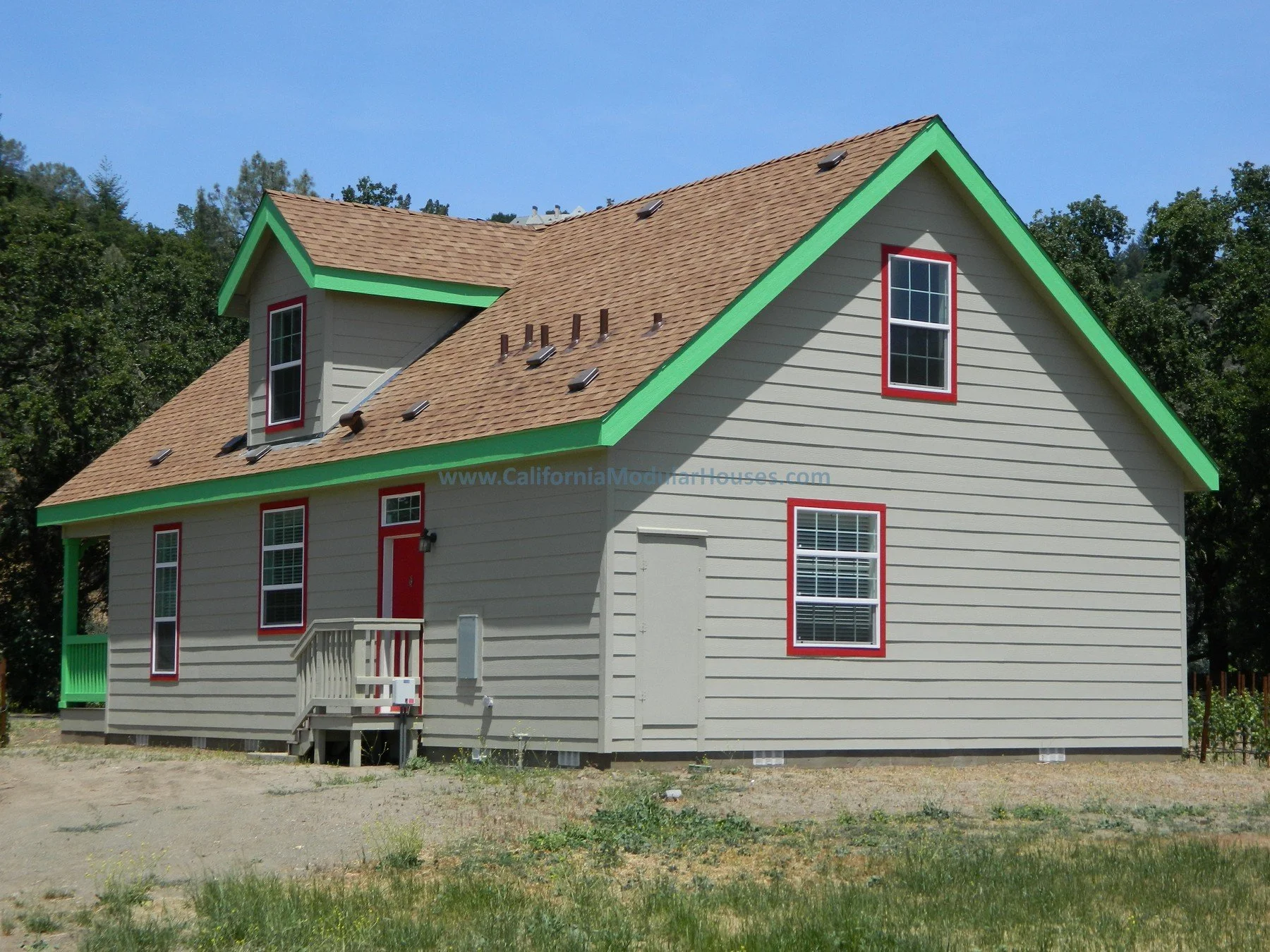 A beige two-story house with green trim on the edges of the roof and red window frames, brown shingle roof, and small front porch, set on a grassy area with trees in the background.