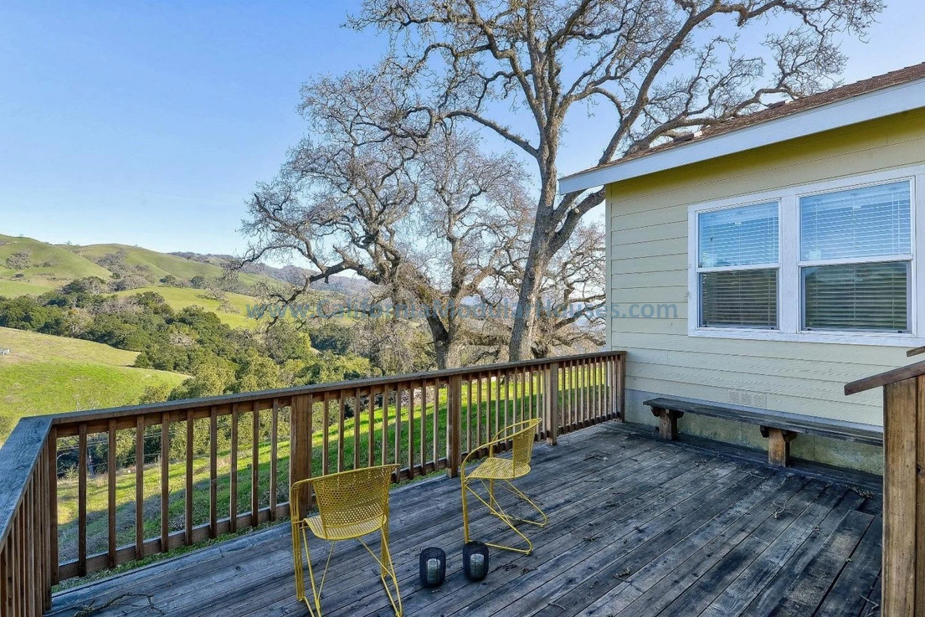 A wooden deck with two yellow chairs and two black lanterns, overlooking green hills and a large leafless tree next to a windowed house. Bay Area Modular Home, Modular Homes California, Modular Homes,