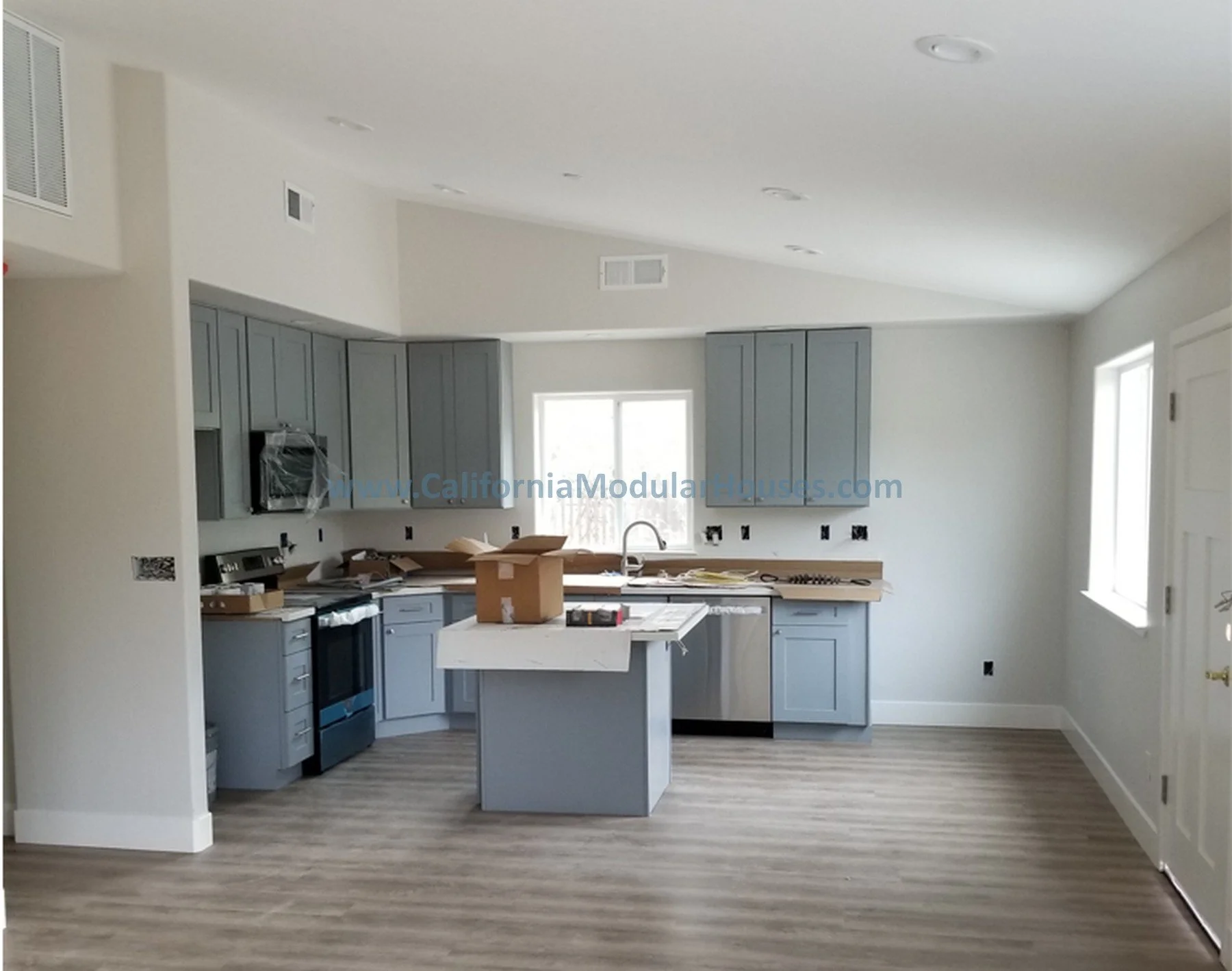 Empty kitchen with gray cabinets, a central island, a window above the sink, and appliances protected in plastic.  Modular granny flat.  California.   