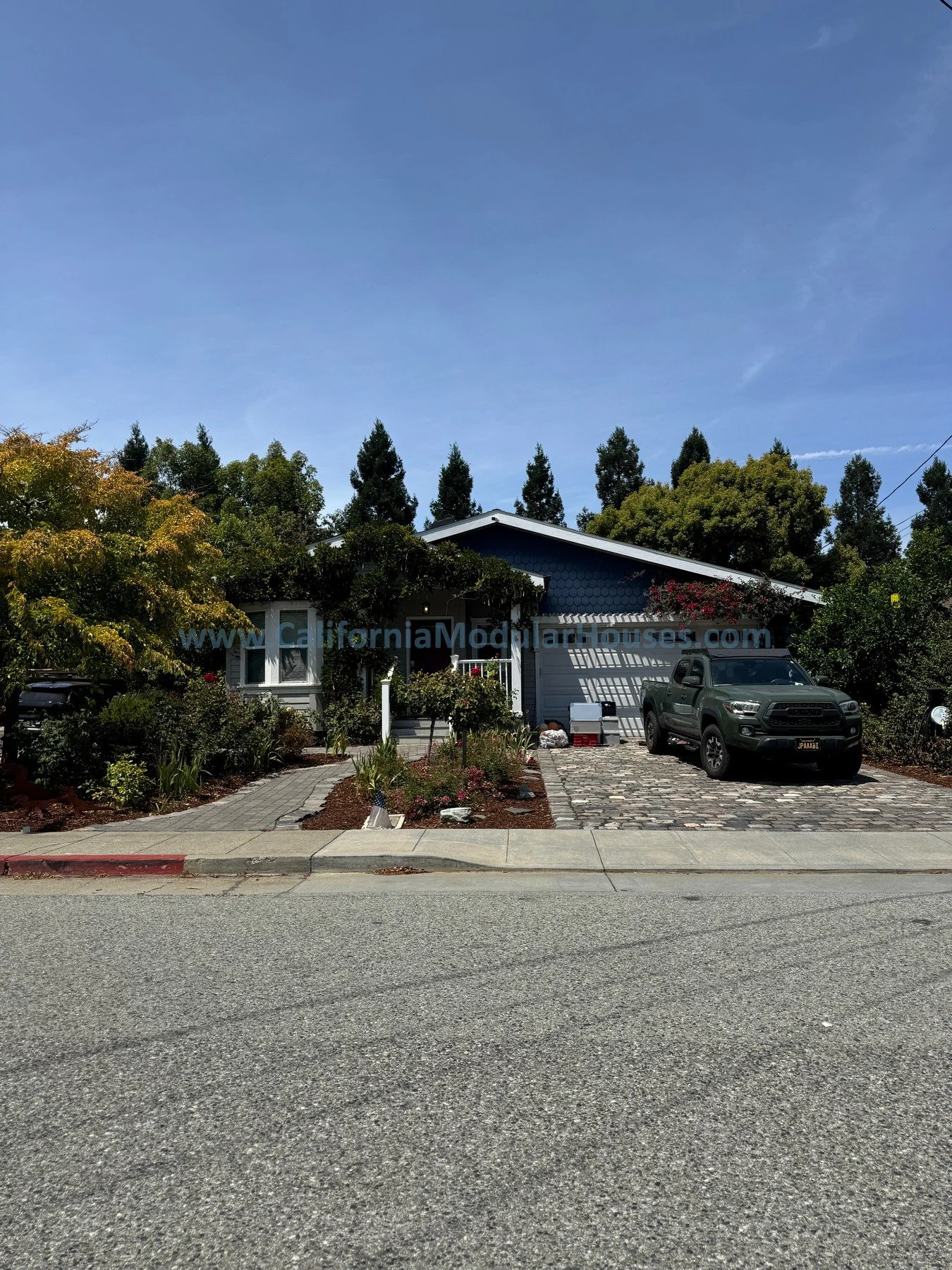 A suburban house with a manicured front yard, a driveway with cobblestones, a black pickup truck parked in the driveway, a blue house with white trim, a garage, surrounded by trees and shrubs, under a clear blue sky.