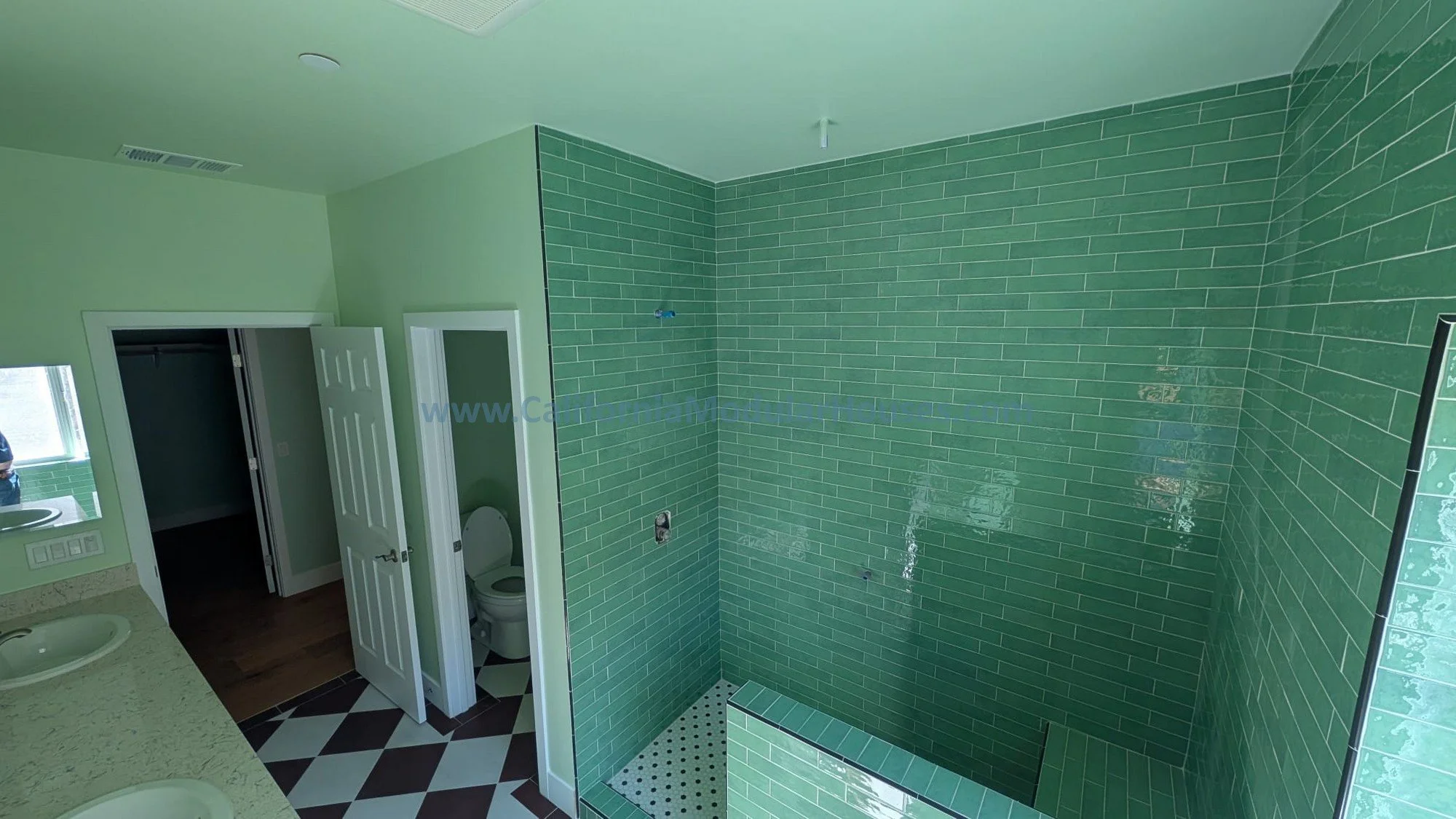 Bathroom with green tiled shower area and a small bench, two white sinks with marble countertops, green walls, and a toilet in a separate small room.