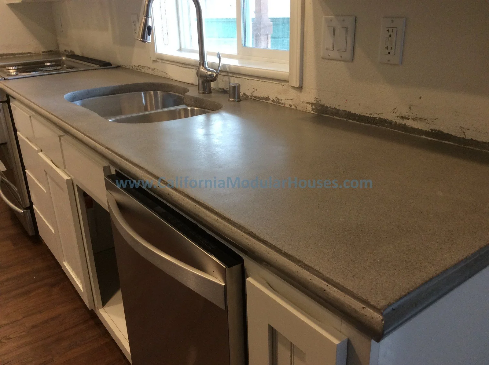 Kitchen countertop with a double sink, faucet, window, and electrical outlets, with a dishwasher and cabinet below.