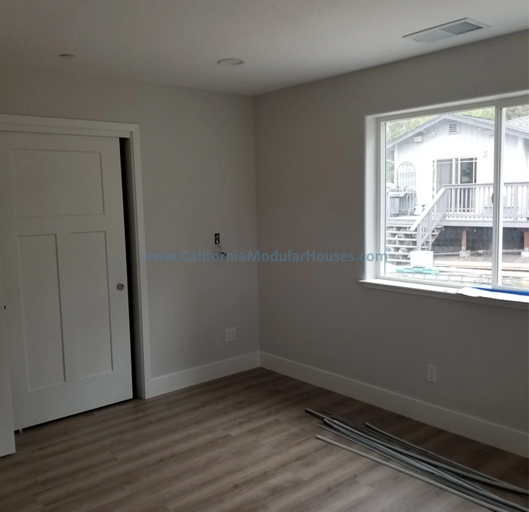 An empty room with a window showing an outdoor deck, a closed closet door, and some rolled-up molding or trim on the wooden floor.  Interior Modular Prefab ADU Photograph