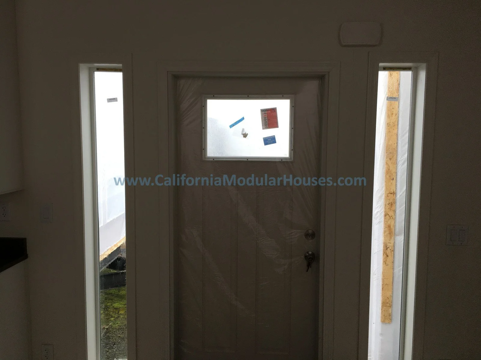 Interior view of a door with a safety viewing window, rectangular, frosted glass window, flanked by two narrow vertical windows, with the website www.CaliforniaModularHouses.com written across the door.  Modular Home.  