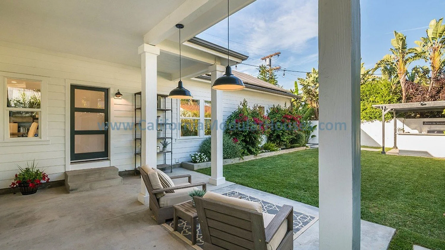 View of a rear covered patio with two chairs, a small table, and hanging pendant lights; a grassy backyard with flowering bushes, trees, a shed, and a white fence in the background.   This is how we custom designed the modular home.  