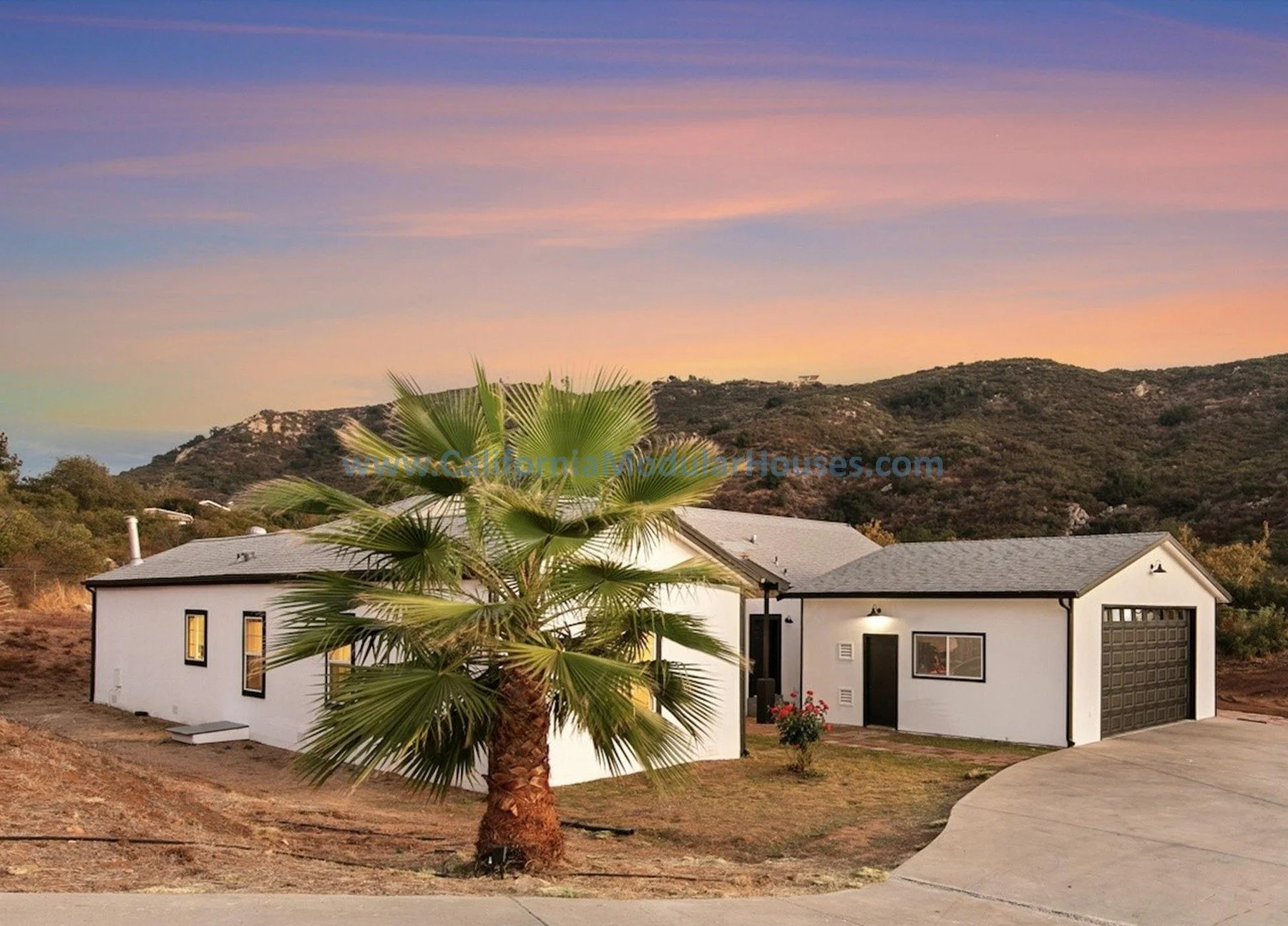 A modern single-story house with a gray roof, white exterior walls, and large windows, situated in a hilly landscape with a palm tree in the front yard during sunset.