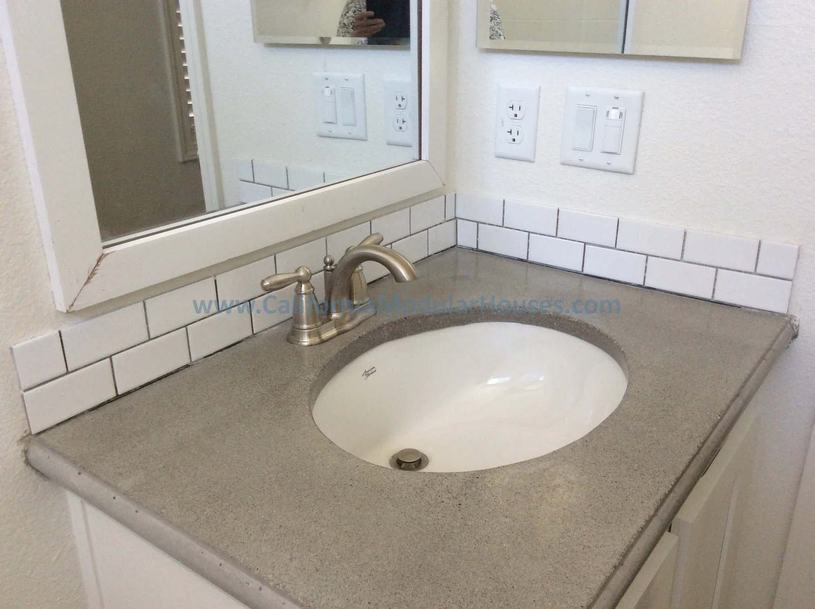 Bathroom sink with a beige countertop, a silver faucet, a large mirror, and white wall tiles with a white textured wall and electrical outlets in the background.
