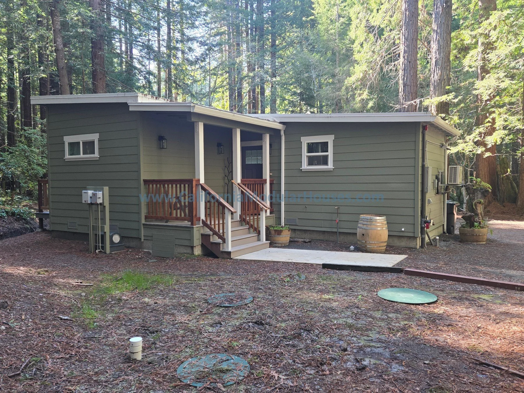 Green single-story house with white trim located in a dense forest, with a small front porch, stairs, and decorative barrels outside. Modular Single Family Residence. Mendocino County, California. CA Modulars