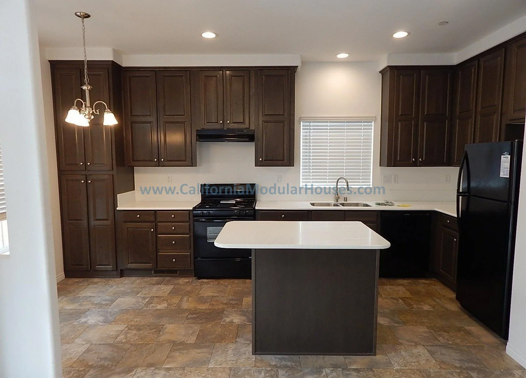 A modern kitchen with dark wood cabinets, a white countertop island, a black stove, a stainless steel sink, and a black refrigerator. There is a window with blinds and recessed ceiling lights, with a pendant light hanging over the island.