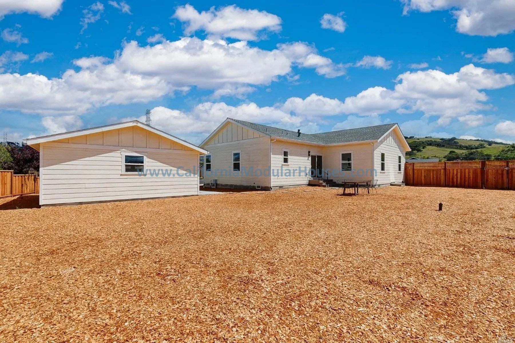 Newly constructed white modular house with a fenced backyard, set against a blue sky with white clouds and green hills in the distance.