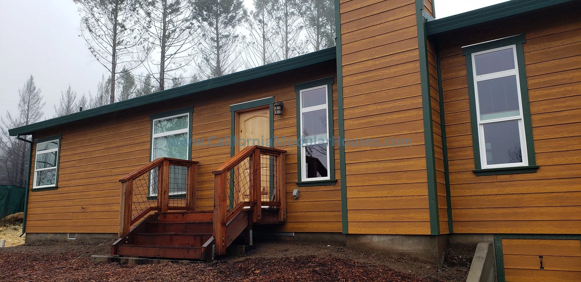 This view shows the intentional rustic look of this Factory Built Home with stairs and a landing to match the stained fiber-cement siding which meets the high fire codes and a site built fire place was installed after the home was delivered.  
