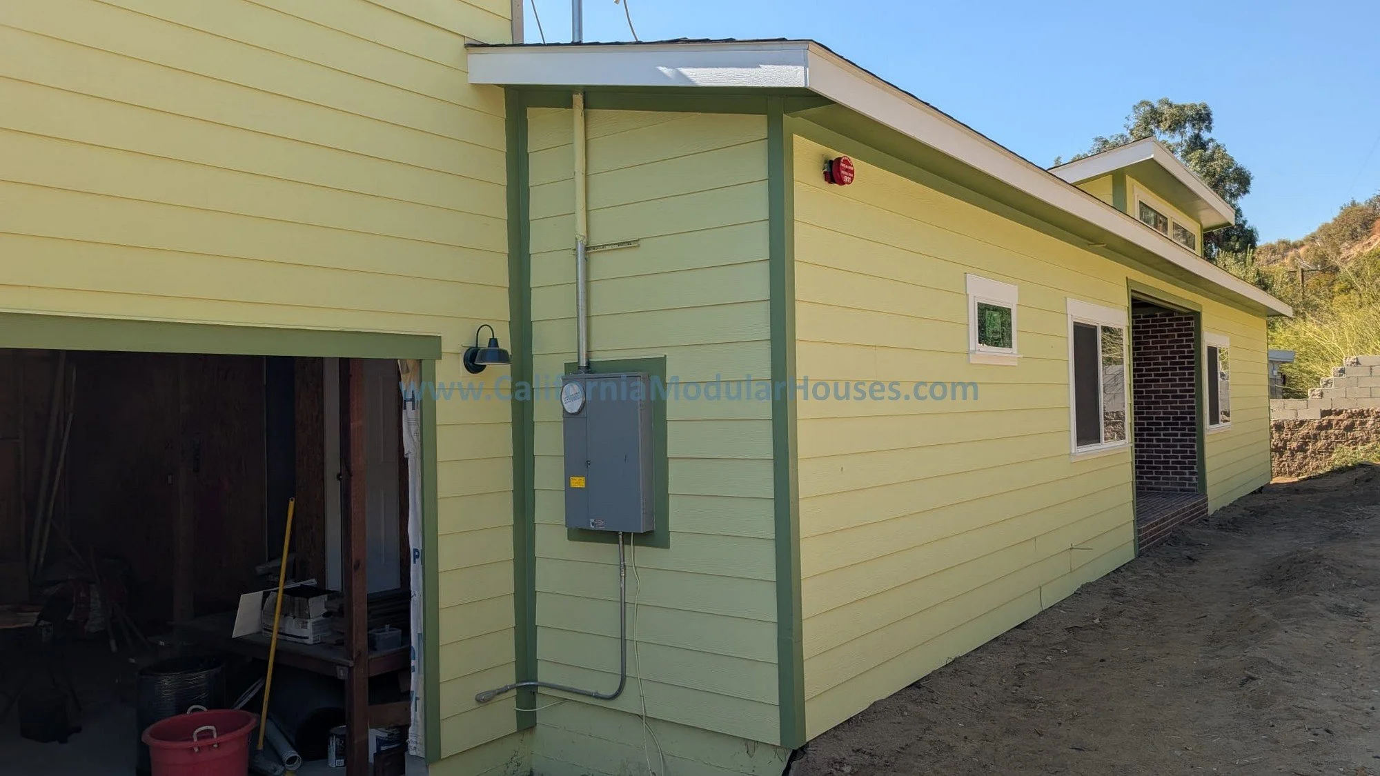 A yellow corner of a house under construction with green trim, two small windows, exposed electrical box, and a partially open garage showing construction supplies inside.