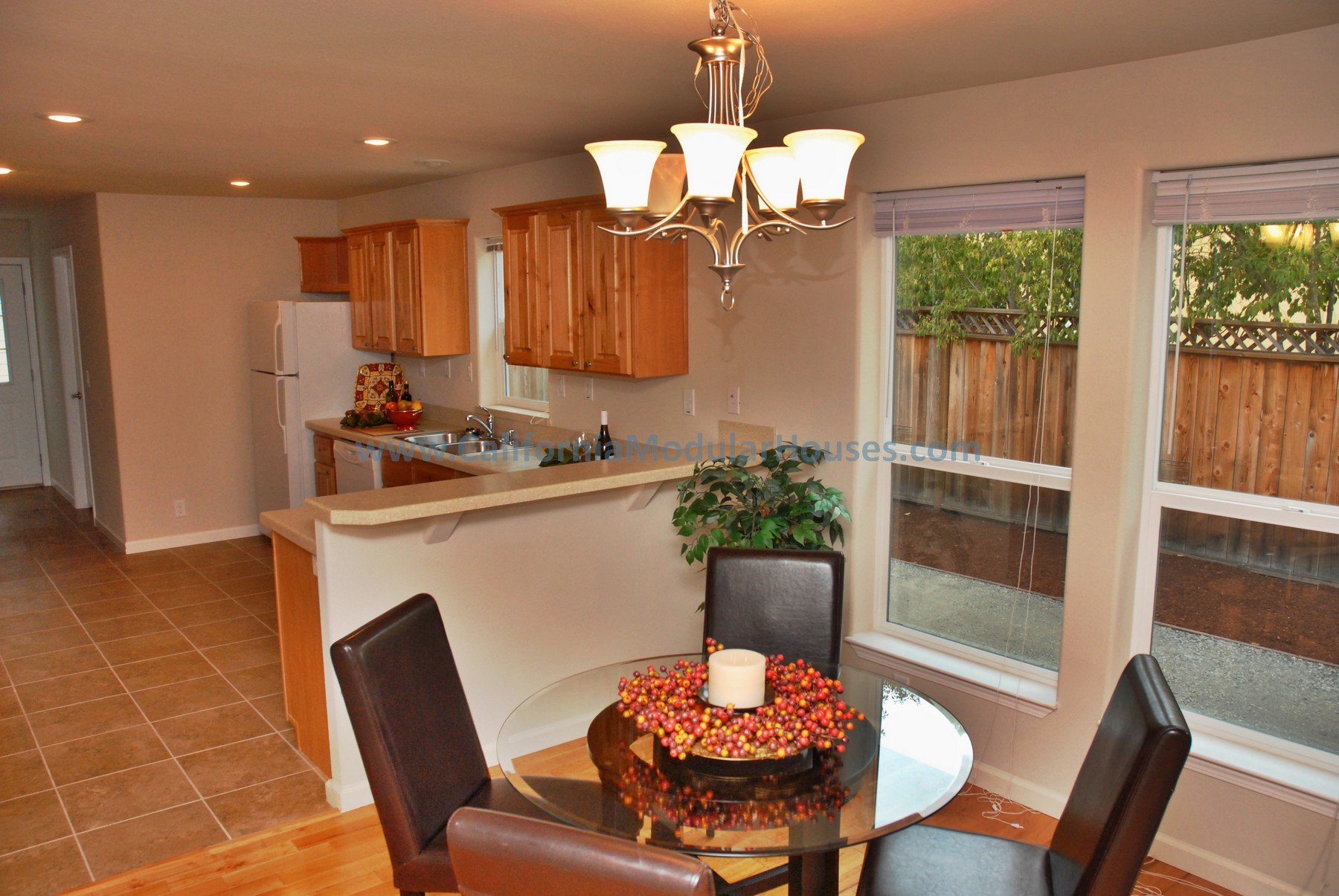 Kitchen and dining area with a round glass table, four black chairs, a chandelier, wooden cabinets, a refrigerator, and large windows letting in natural light.