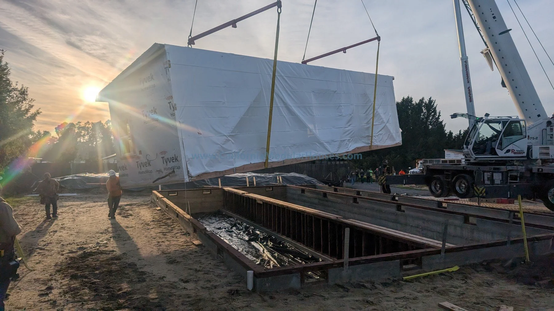 A large construction site during sunset shows a crane lifting a prefabricated building wrapped in white plastic. Several workers are present, and there is a deep rectangular excavation with reinforced sides in the foreground.  Modular prefab ADU.  
