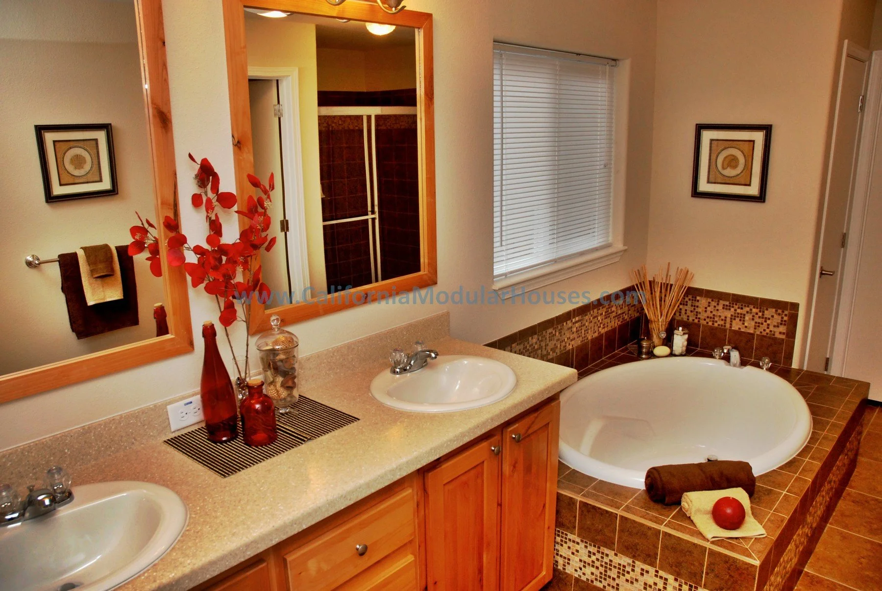 Bathroom with double sinks, a large mirror, decorative vases, a window with blinds, and a built-in bathtub with brown tiles and a rolled towel, an apple, and bath accessories placed on the tub's ledge.