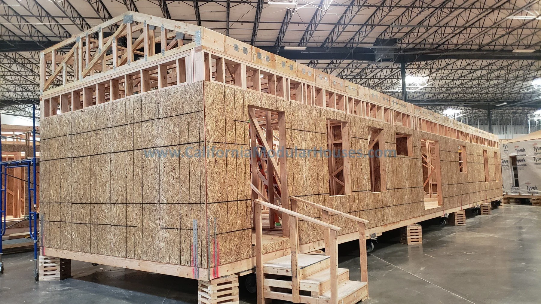 Construction of a building in an indoor warehouse with wooden framing, oriented strand board exterior walls, small stairs leading to the entrance, and a steel roof structure overhead.
