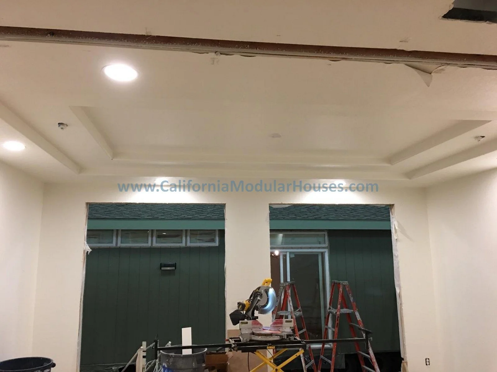 Interior of a room being build with two green doors, a circular saw on a worktable, ladders, and construction tools, with a website address on the wall.  New House.  