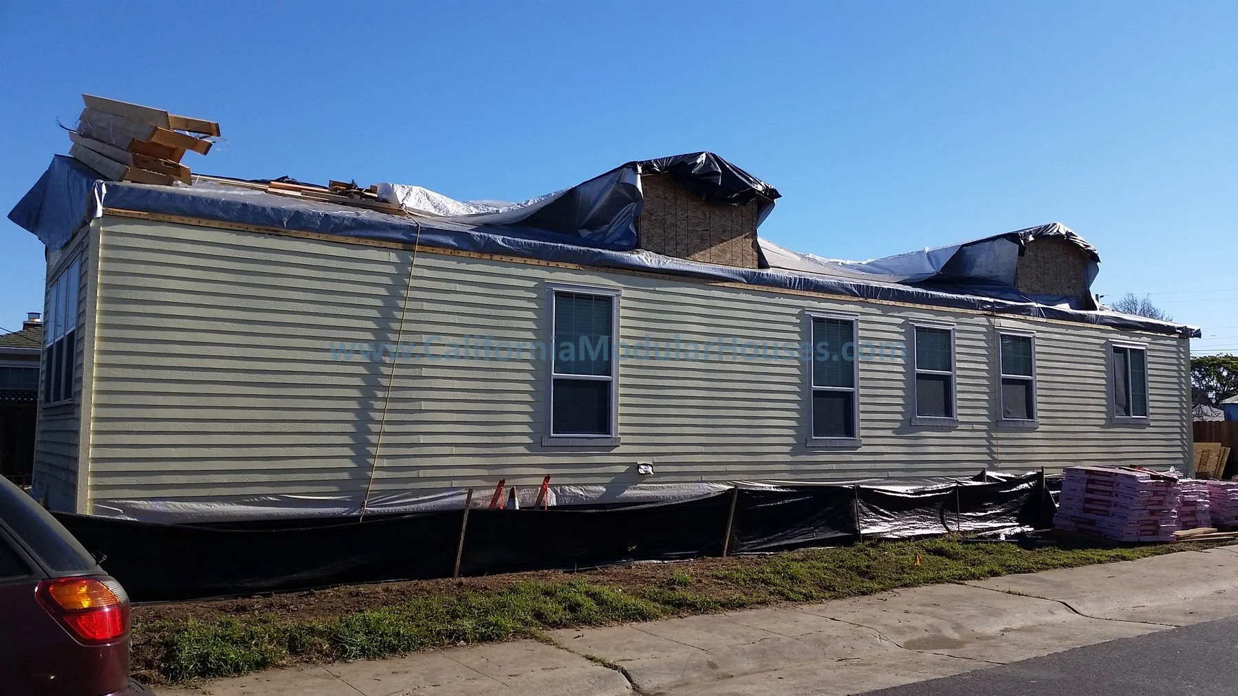 A house under construction with siding, windows, and a temporary roof covering, surrounded by construction materials and a black barrier. Bay Area Modular Home, Modular Homes in California,