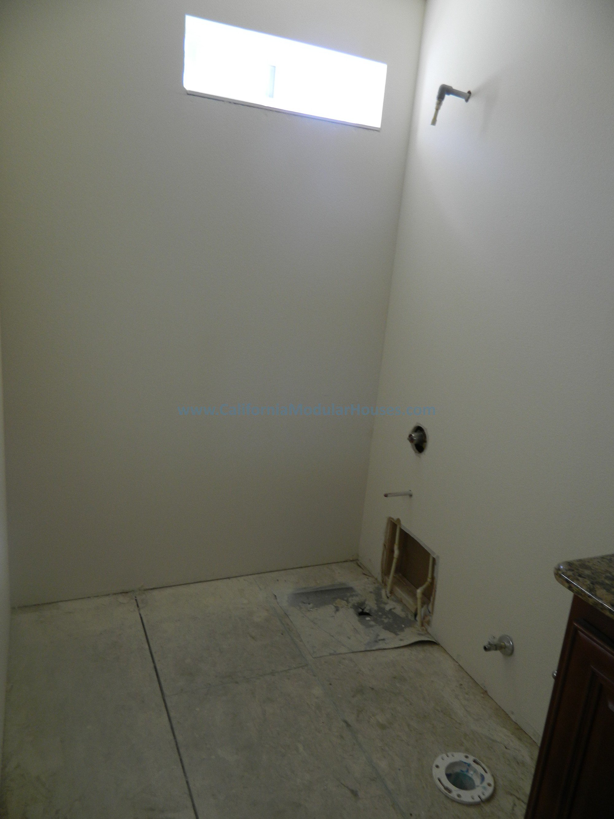 An unfinished laundry room or bathroom with exposed plumbing and electrical fixtures, a small window near the ceiling, and a partially open wall access panel on the floor.