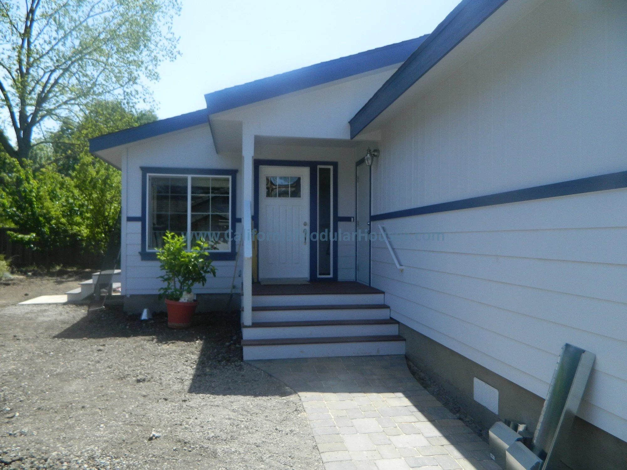 Newly constructed house with white and blue exterior, front steps leading to the door, potted plant at the entrance, and partial view of a paved walkway.