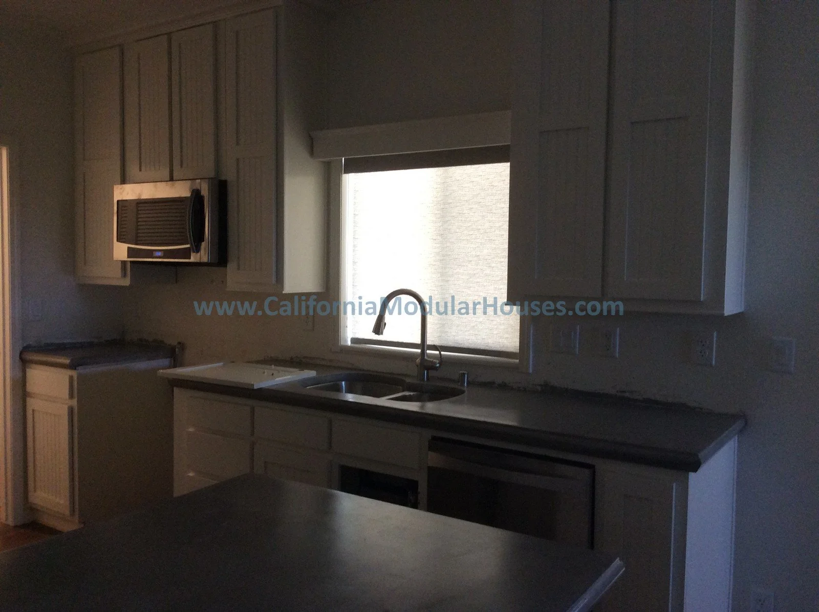 Kitchen with beige cabinets, a stainless steel microwave, a double sink with a window above, and the website www.CaliforniaModularHouses.com printed across the window.