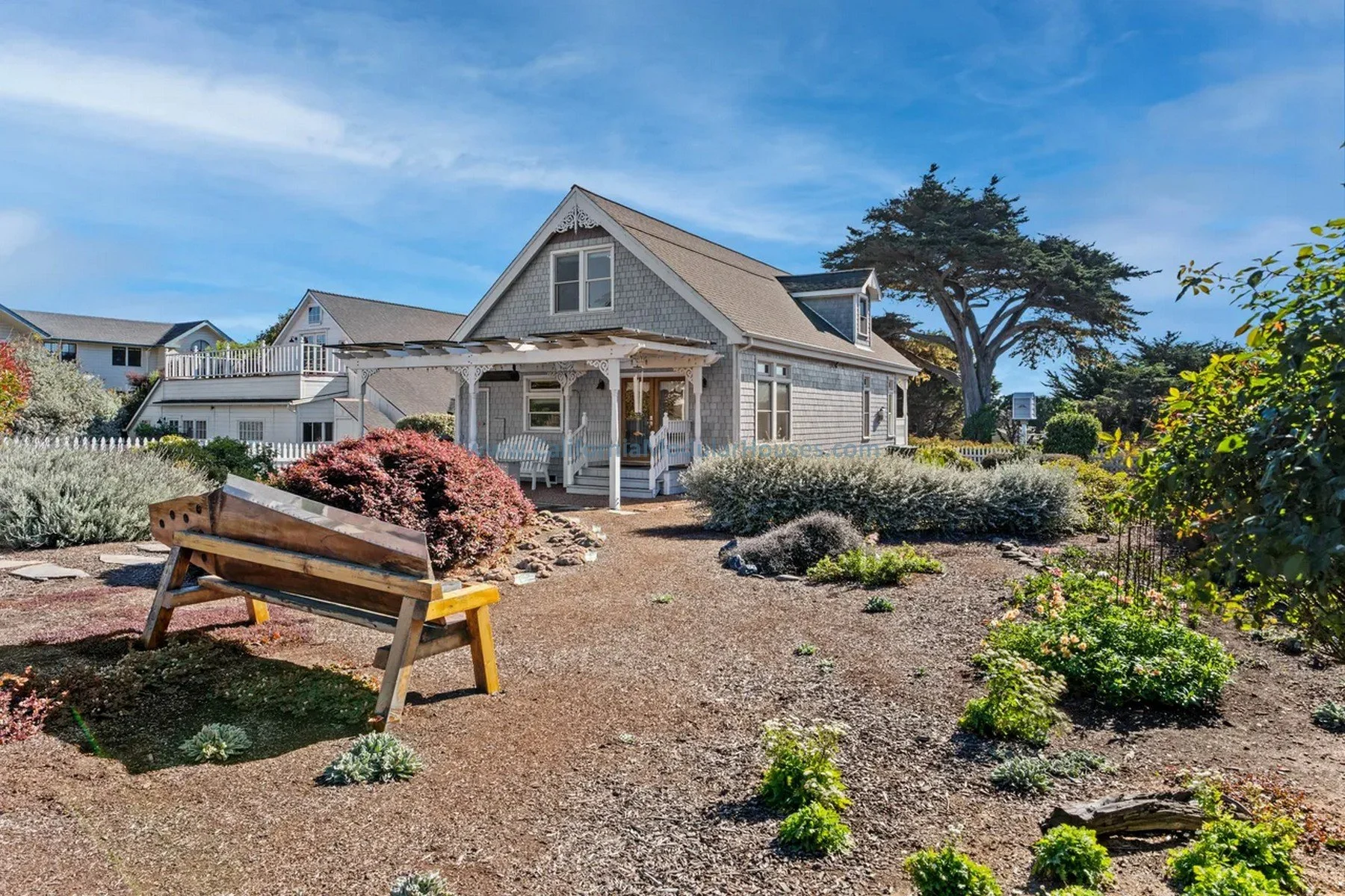 A house with gray shingle siding, a porch with white railings, and a front yard with a garden and a wooden bench on a dirt pathway. California. Pre-fab modulars. Custom design coastal commission cape cod modular home.