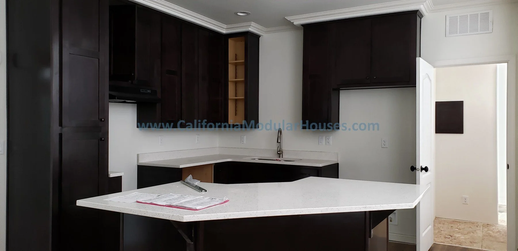 Kitchen with dark cabinets, white countertop, and a sink with a faucet. There is an open space for a stove and electrical outlets on the wall. A door is open to a tiled room or hallway. The kitchen is being completed in the factory.  Modular home.  