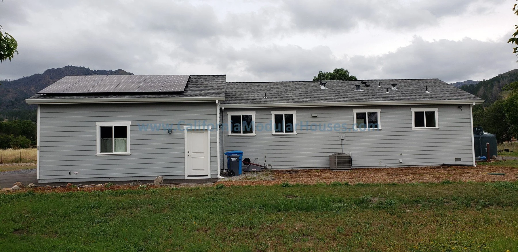Side view of a gray, one-story modular ADU with five windows, a door, and solar panels on the roof, set in a landscape with mountains in the background and a cloudy sky overhead.  Prefab Sonoma County, CA