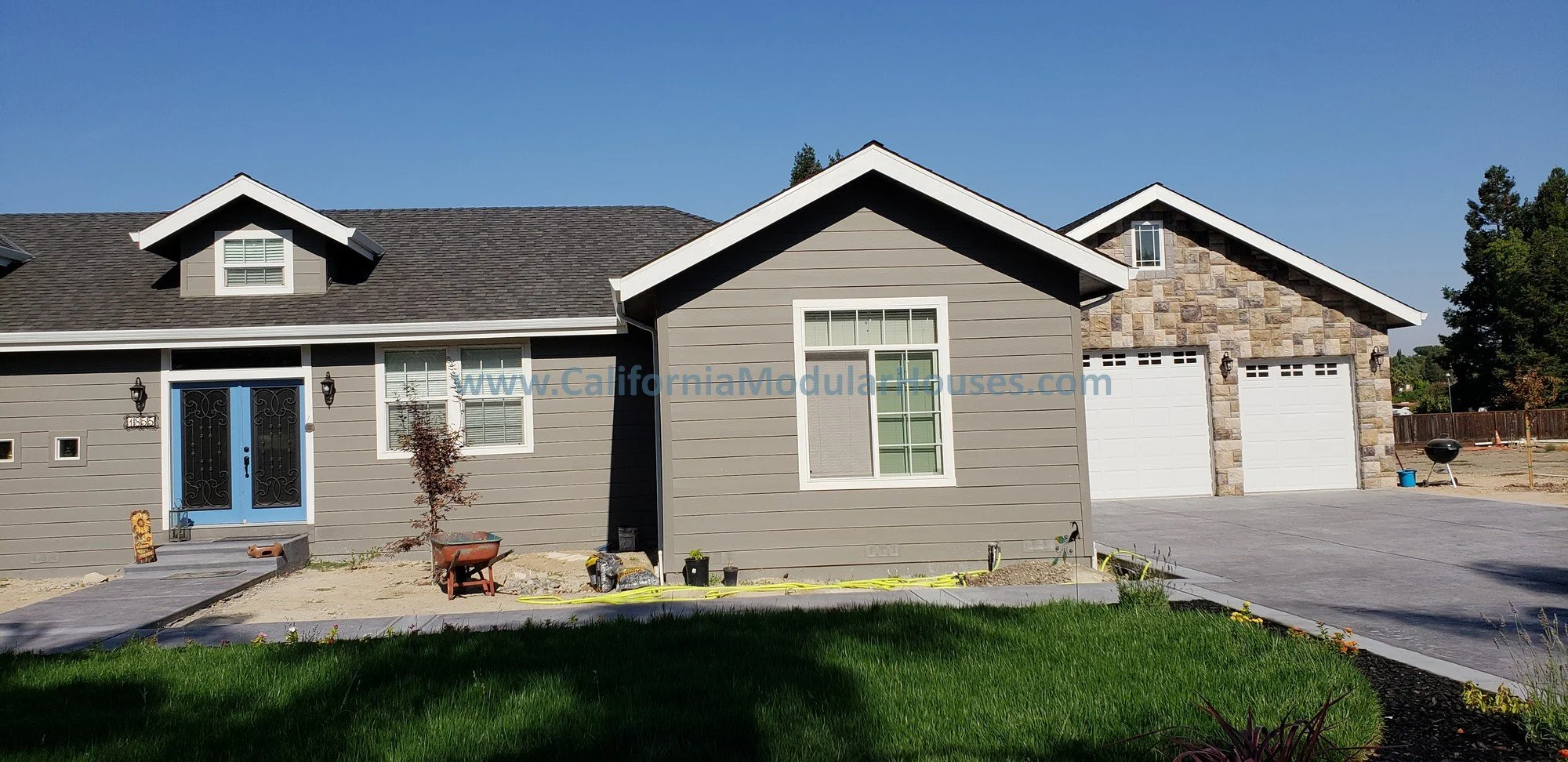 Front of a new house with grey siding, white trim, and a stone accent wall. It has a double garage and a small front yard with green grass, a small tree.   This modular home with a large kitchen is in Contra Costa County, California.  