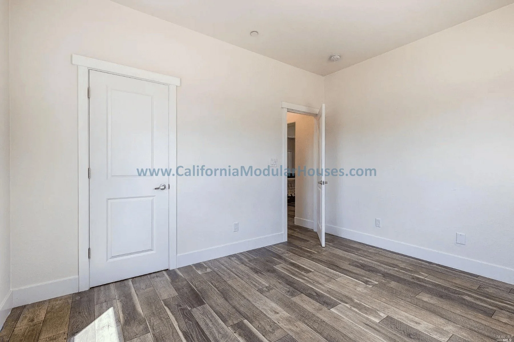 Empty room with white walls, wood flooring, a closed white door on the left, an open door in the middle, and no furniture.