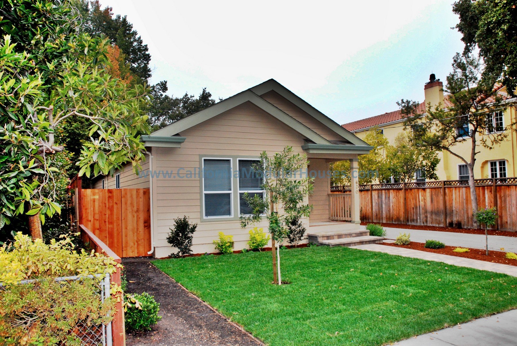 A small beige house with a gable roof, front porch steps, and a green lawn with young trees and bushes, surrounded by a wooden fence.