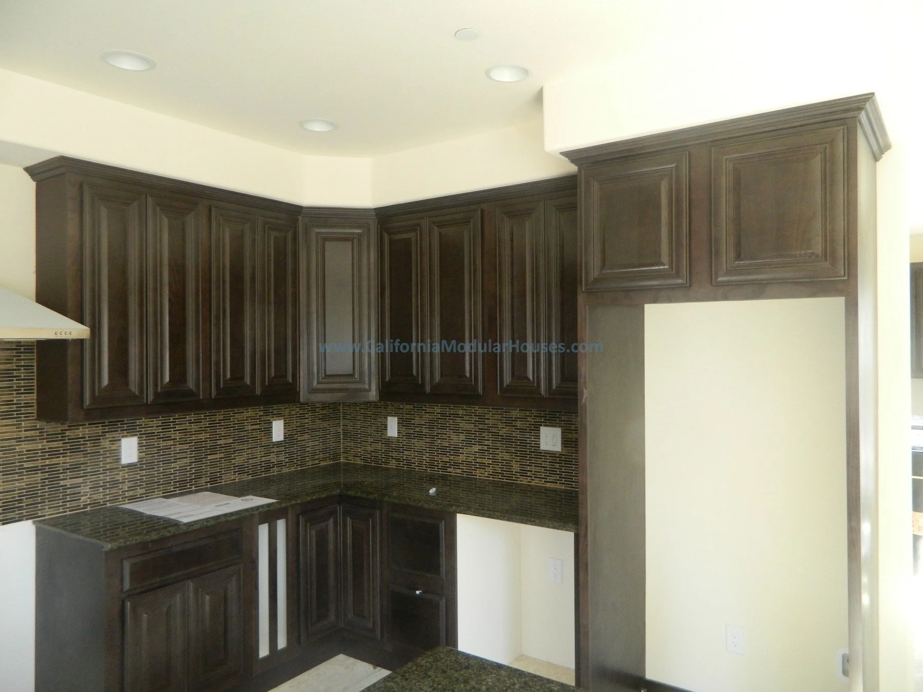 Kitchen with dark wooden cabinets, granite countertops, mosaic tile backsplash, and an empty space for a refrigerator.  Modular Homes in California, Modular Homes California, Modular Homes, California Modular.