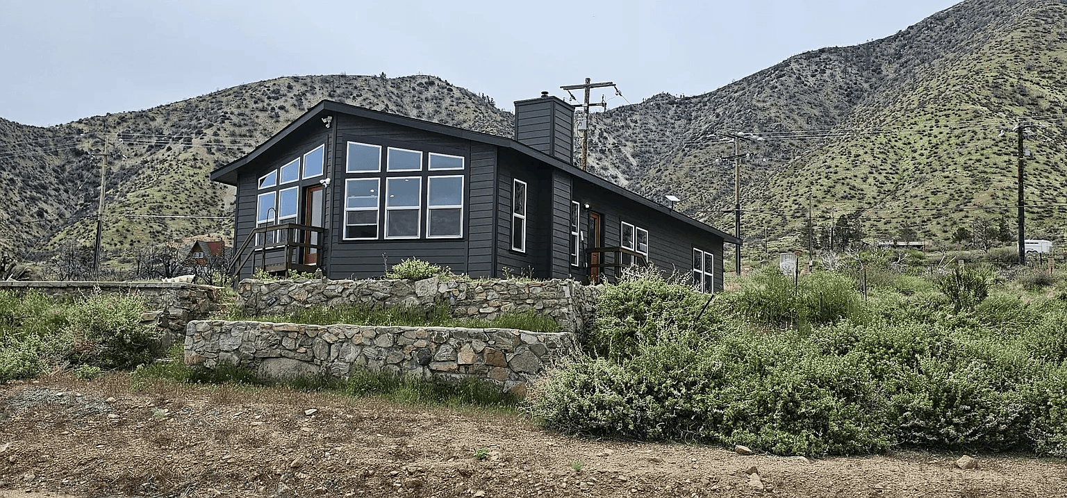 Newly constructed yellow house with multiple doors and windows, a paved backyard, and trees with fall foliage in the background.