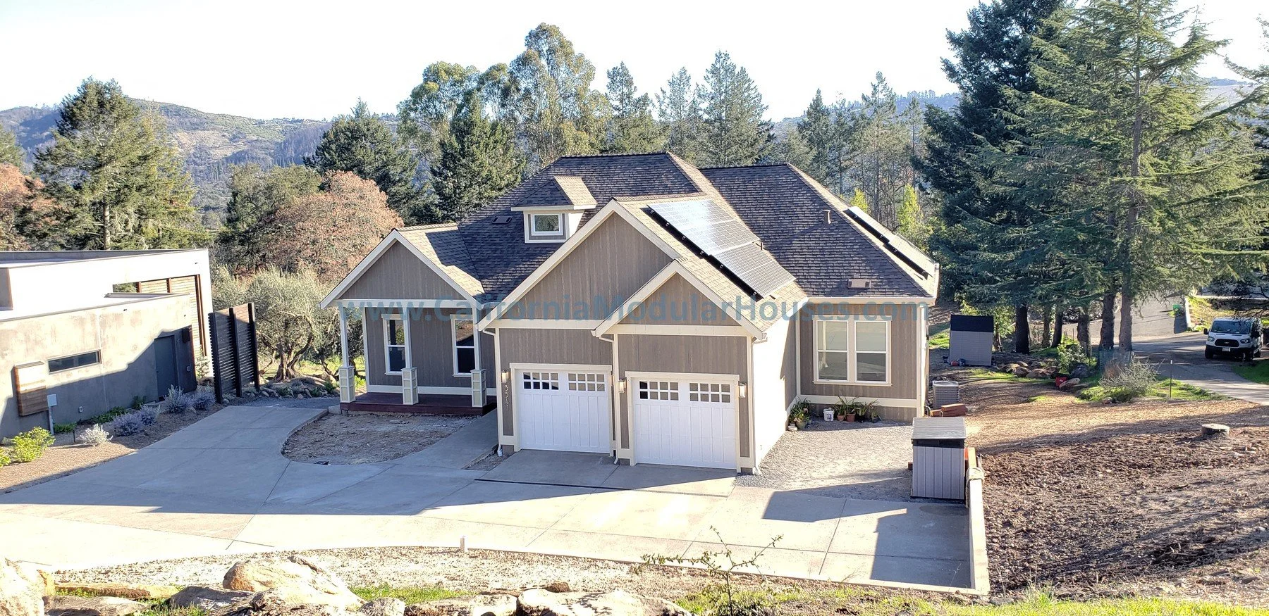New gray house with white garage doors, solar panels on the roof, large trees in the background, and a driveway.