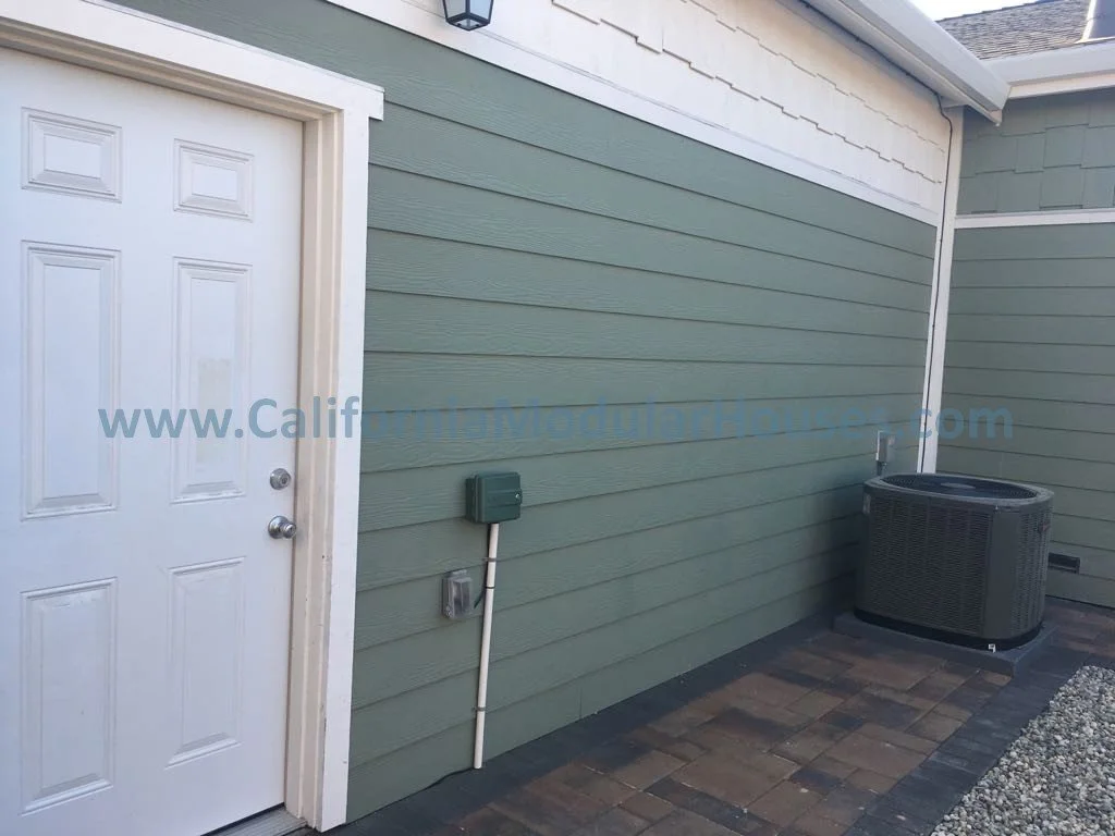 Exterior view of a house with green siding, a white door, and an air conditioning unit outside. There is also a small electrical box on the wall.
