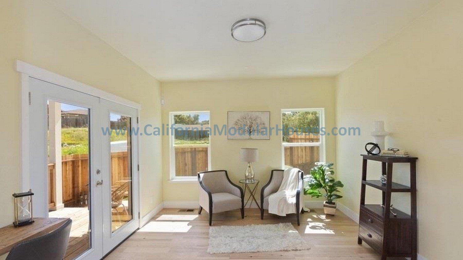 Sunlit living room with two chairs, a small side table with a lamp, a potted plant, and a wooden shelf against the wall, with large windows and a glass door leading outside.   San Francisco Bay Area, CA.   California modular home.  prefab modular CA